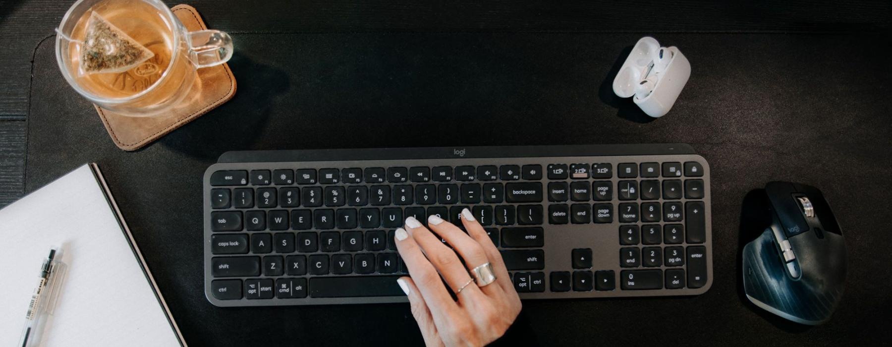 woman's hand on a keyboard surrounded by office items and a cup of tea on a coaster